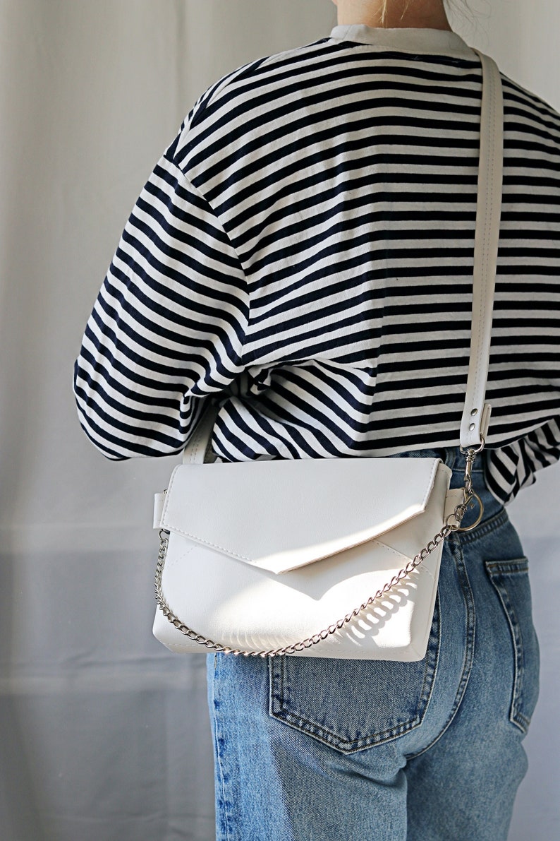 white bag on woman's shoulder with a silver chain envelope bag minimalistic design Vegan leather bag small handbag mini shoulder bag blue stripes and light blue jeans.