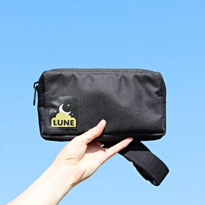 Puede incluir: Riñonera negra con cierre de cremallera y correa ajustable. La riñonera tiene un logotipo dorado con una luna creciente y estrellas y la palabra "LUNE" impresa en ella.