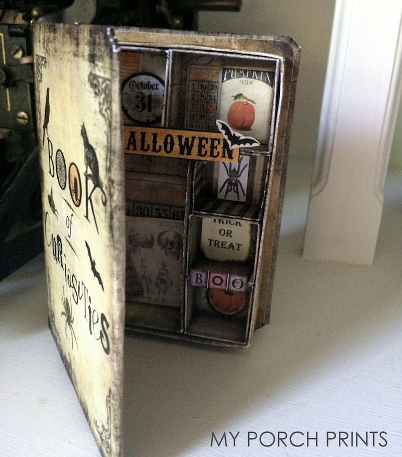 Puede incluir: Un libro de estilo vintage titulado "Book of Curiosities" con tem&aacute;tica de Halloween. El libro est&aacute; abierto para revelar una serie de peque&ntilde;os compartimentos llenos de decoraciones de Halloween, incluyendo un calendario, una calabaza, una ara&ntilde;a y un letrero de "Trick or Treat".