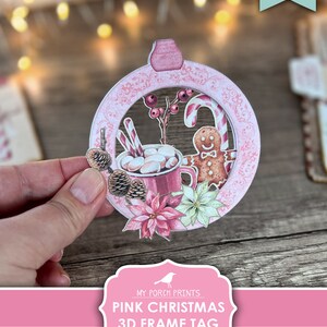 Puede incluir: Etiqueta de marco 3D de Navidad rosa imprimible. La etiqueta presenta un c&iacute;rculo rosa con un borde blanco y una escena de invierno en su interior. La escena incluye una taza de chocolate caliente con malvaviscos, un hombre de jengibre, bastones de caramelo y poinsettias.