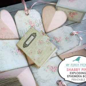 May include: A printable shabby pink exploding ephemera box with a vintage floral design. The box is made of paper and has a heart-shaped lid. The box is decorated with ribbons, tags, and a small notebook. The box is perfect for storing small items or gifts.