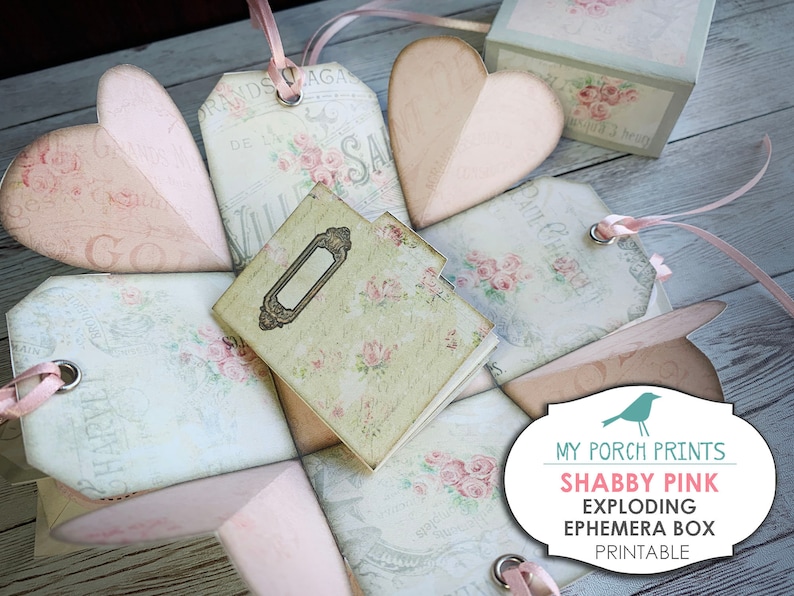 May include: A printable shabby pink exploding ephemera box with a vintage floral design. The box is made of paper and has a heart-shaped lid. The box is decorated with ribbons, tags, and a small notebook. The box is perfect for storing small items or gifts.