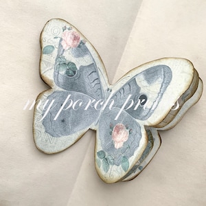 Puede incluir: Una decoraci&oacute;n artesanal de mariposa tridimensional en capas, con un dise&ntilde;o floral vintage. La mariposa est&aacute; hecha de papel y tiene un esquema de color azul claro y blanco con rosas rosas.