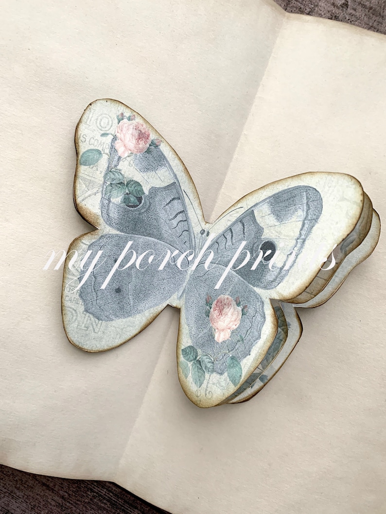 Puede incluir: Una decoraci&oacute;n artesanal de mariposa tridimensional en capas, con un dise&ntilde;o floral vintage. La mariposa est&aacute; hecha de papel y tiene un esquema de color azul claro y blanco con rosas rosas.