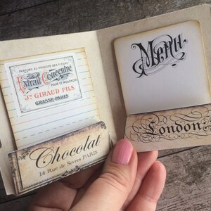 May include: A vintage-style menu card with a handwritten "Menu" title and a decorative "London" label. The card is open to reveal a small, rectangular label with the text "Chocolat 14 Rue de Sevres PARIS" and a smaller label with the text "PARFUMS AU PRINCIPE DES FLEURS Extrait Concentre POUR LE MOUCHOIR J. GIRAUD FILS GRASSE PARIS".