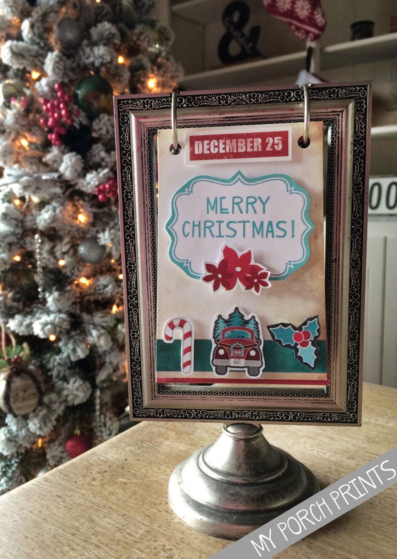 May include: A silver framed picture with a white background and the text "DECEMBER 25" and "MERRY CHRISTMAS!" in red and blue lettering. The picture also features a red truck with a Christmas tree on top, a candy cane, and a holly sprig.