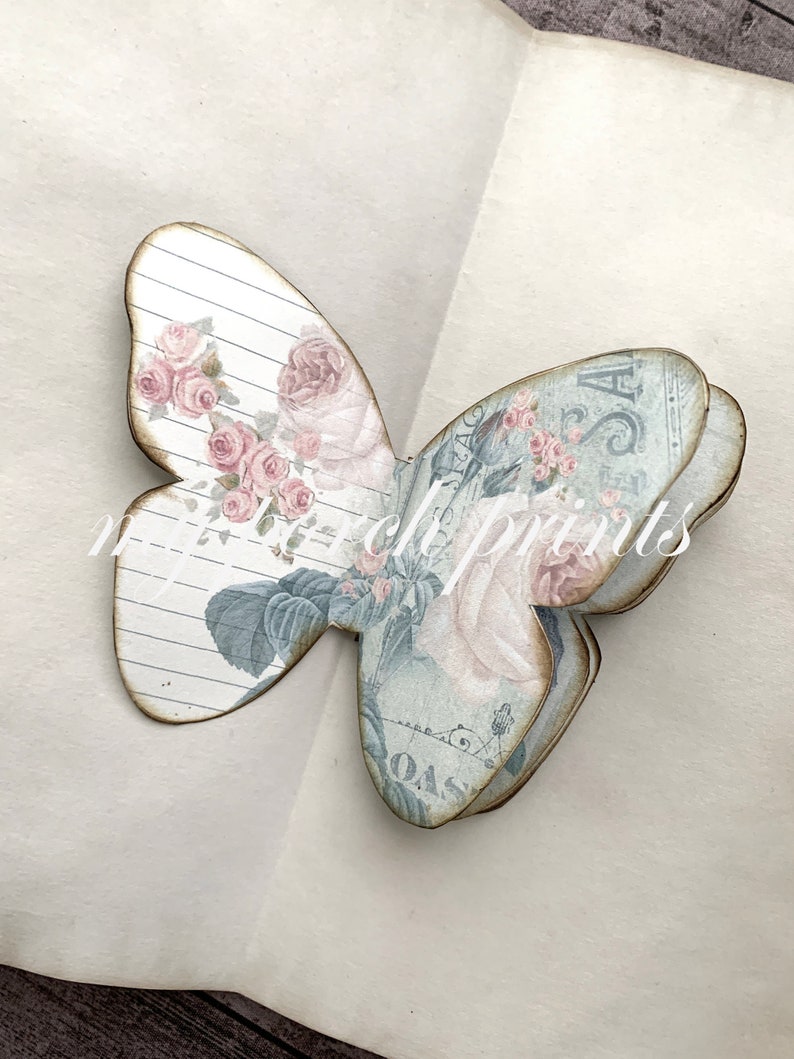 Puede incluir: Una mariposa de papel en capas con un dise&ntilde;o floral vintage. La mariposa est&aacute; hecha de m&uacute;ltiples capas de papel, cada una con un patr&oacute;n diferente. La mariposa es de color azul claro y rosa con un fondo blanco.