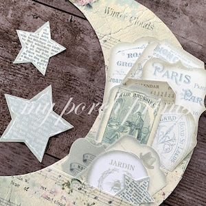May include: A paper moon and stars collage with vintage ephemera. The moon shape features text, musical notes, and a floral design. The collage includes labels with text like "Paris" and "Jardin".