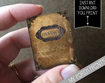 Puede incluir: Una descarga digital imprimible de una portada de libro de estilo vintage con fondo dorado y borde adornado. La portada presenta una placa de metal con el texto "DA VINCI" en letras negras.
