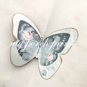 Puede incluir: Una mariposa de madera decorativa con un dise&ntilde;o vintage que presenta una mariposa gris con rosas rosadas y hojas verdes. La mariposa est&aacute; sobre un fondo blanco.