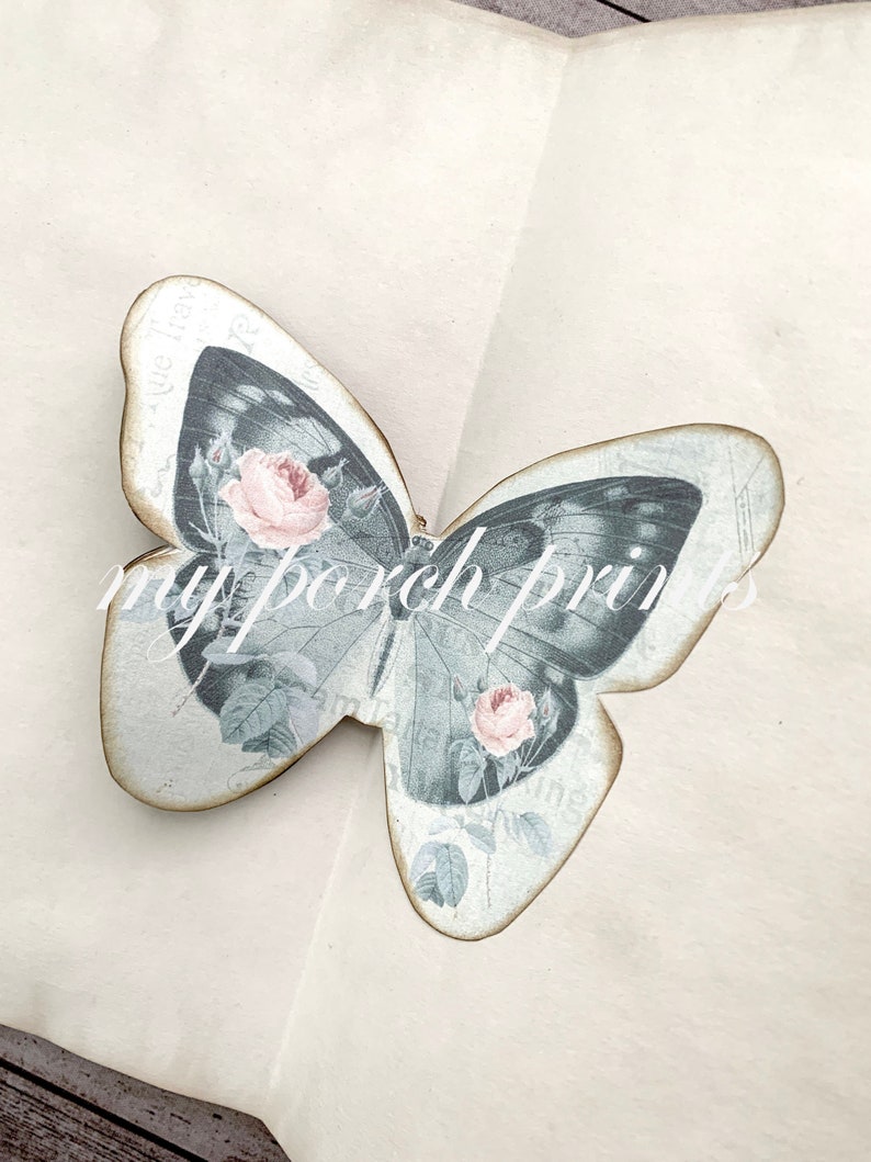 Puede incluir: Una mariposa de madera decorativa con un dise&ntilde;o vintage que presenta una mariposa gris con rosas rosadas y hojas verdes. La mariposa est&aacute; sobre un fondo blanco.