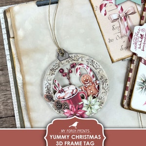 May include: Printable 3D Christmas frame tag with a red mug of hot chocolate, candy canes, a gingerbread man, and poinsettias. The tag is decorated with a white and gold ornate frame. "Yummy Christmas 3D Frame Tag Printable" is written on the tag.