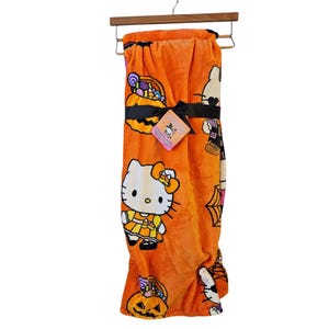 Könnte beinhalten: Eine orangefarbene Fleece-Decke mit Halloween-Motiv, auf der Hello Kitty in einem Hexenkostüm zu sehen ist. Die Decke hat ein schwarzes Band mit einem Hello Kitty Halloween-Etikett.