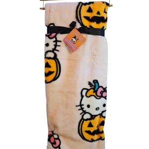 Könnte beinhalten: Eine Plüschdecke mit Hello Kitty, die als Kürbis verkleidet ist. Die Decke ist cremefarben mit orangefarbenen und schwarzen Akzenten. Die Decke hat den Text "Hello Kitty Halloween" auf dem Etikett.