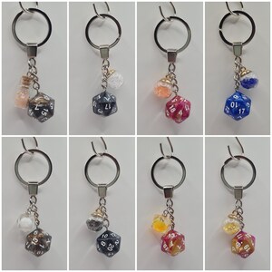 Pode incluir: Seis chaveiros diferentes com um aro de chave prateado e um pequeno frasco de vidro com glitter e um dado de 20 faces. Os dados são de diferentes cores: preto, vermelho, azul, cinza, amarelo e laranja.