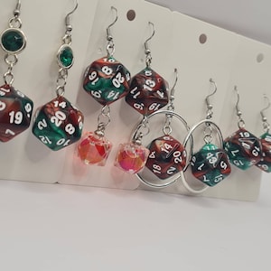 Könnte beinhalten: Ein Set mit sechs Hängeohrringen mit roten, grünen und schwarzen Würfeln mit weißen Zahlen. Die Ohrringe sind aus silberfarbenem Metall gefertigt und haben ein Ringdesign.