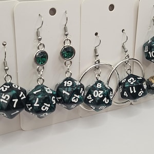 Op de afbeelding: Een set hangende oorbellen met groene en zilveren dobbelsteenbedels. De dobbelstenen zijn genummerd met witte cijfers. Sommige oorbellen hebben een klein groen steentje bovenaan de hanger.