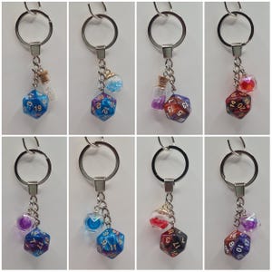 May include: Set of eight keychains featuring a 20-sided die with a different colour and glitter combination, each with a small glass vial charm. The vials are filled with glitter and have a cork stopper. The keychains are silver.