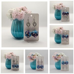 Könnte beinhalten: Ein Paar Hängeohrringe mit einem silbernen Anhänger und einem blauen und violetten Würfelanhänger. Die Ohrringe werden auf einer weißen Karte mit einem floralen Hintergrund präsentiert.