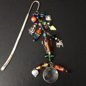 Puede incluir: Un marcapáginas plateado con un mango largo y delgado y un amuleto colorido que cuelga del extremo. El amuleto presenta una variedad de cuentas, incluyendo naranja, verde, azul y plateado. También hay pequeños amuletos plateados en forma de corazones, perros y una lupa.
