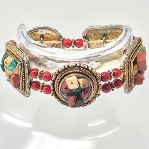 May include: A multi-strand bracelet with red coral beads and three decorative elements. Each element is a circular frame with a variety of colorful stones, including green, red, and brown.