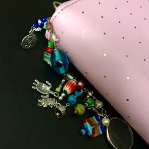 May include: A pink zippered pouch with a colorful beaded charm. The charm features a blue evil eye bead, a red and white striped bead, a silver bell, and a silver seahorse charm.