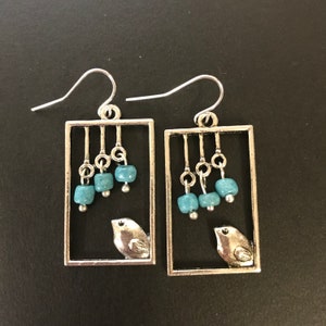 May include: Silver earrings with a rectangular frame and a small bird charm. The frame has a dangling design with turquoise beads.