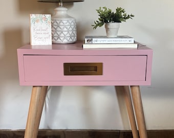 May include: A pink nightstand with a drawer and brass-colored handle. The nightstand has light-colored wooden legs. A lamp with a gray shade, a decorative vase, a small potted plant, and books are on top. A card with text is also on top.