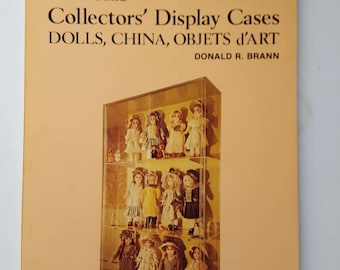 May include: A vintage book cover with the title "how to build Collectors' Display Cases" featuring a glass display case filled with dolls. The book is titled "DOLLS, CHINA, OBJETS d'ART" by Donald R. Brann. The cover has a light orange background.