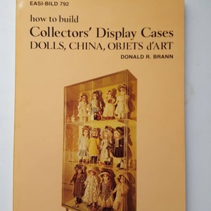 May include: A vintage book cover with the title "how to build Collectors' Display Cases" featuring a glass display case filled with dolls. The book is titled "DOLLS, CHINA, OBJETS d'ART" by Donald R. Brann. The cover has a light orange background.