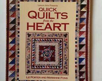 以下が含まれることがあります： Liz Porter と Marianne Fons による「Quick Quilts from the Heart」の表紙。 赤、青、クリーム色のパッチワークの三角形のボーダーと、中央にキルトのイラストが描かれたベージュの背景が特徴です。
