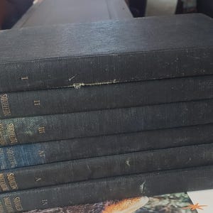 Puede incluir: Una pila de seis libros antiguos con cubiertas azul oscuro. Las lomos muestran letras doradas con títulos como "Course in Modern Production Methods" y "Business Training Corporation". Se ven los números romanos I a VI.