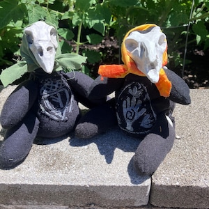May include: Two plush dolls with black fabric bodies and white skull-shaped heads. The left doll wears a green headscarf and features an acorn symbol on its chest. The right doll wears an orange and yellow headscarf and features a hand symbol on its chest.