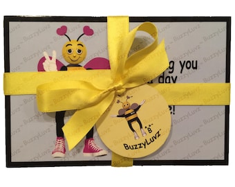 May include: A rectangular gift box wrapped in a bright yellow satin ribbon with a matching circular tag. The box features a cartoon bee character wearing pink sneakers and making a peace sign, with the text BuzzyLuvz printed across the surface.
