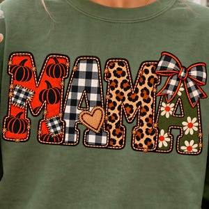 May include: An olive green long-sleeve sweatshirt featuring the word MAMA in large letters. The letters are filled with various patterns including orange pumpkins, black and white buffalo plaid, leopard print, and a green floral design with a bow.