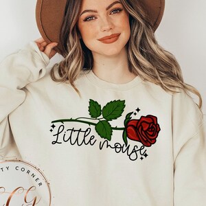 May include: A cream-colored sweatshirt with a red rose and the text "Little mouse" printed on the front. The sweatshirt is being worn by a person with long blonde hair and a brown hat.