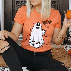 K&ouml;nnte beinhalten: Eine Frau tr&auml;gt einen schwarzen Hut mit wei&szlig;en Sternen und ein pfirsichfarbenes T-Shirt mit einem Geistermotiv, das Ballons h&auml;lt. Der Geist ist wei&szlig; mit einem schwarzen L&auml;cheln und schwarzen Augen. Die Frau sitzt auf einem Teppich mit K&uuml;rbissen im Hintergrund.