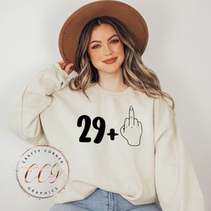 Puede incluir: Una sudadera blanca con el texto "29+" y una ilustraci&oacute;n en blanco y negro de un dedo medio.