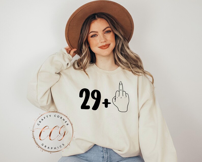 Puede incluir: Una sudadera blanca con el texto "29+" y una ilustraci&oacute;n en blanco y negro de un dedo medio.