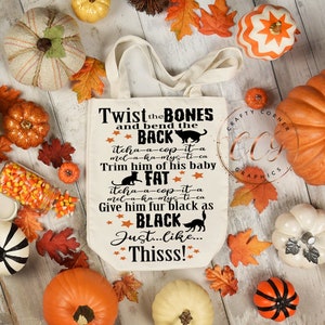 May include: A white canvas tote bag with a black and orange Halloween-themed design. The text on the bag reads "Twist the BONES and bend the BACK itcha-a-cop-it-a mel-a-ka-mys-ti-ca Trim him of his baby FAT itcha-a-cop-it-a mel-a-ka-myc ti-ca Give him fur black as BLACK Just...Like...Thisss!"