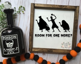 May include: A wooden sign with a black silhouette of three ghosts and the text "Room for one more?" The sign has a distressed white background and a brown wooden frame.