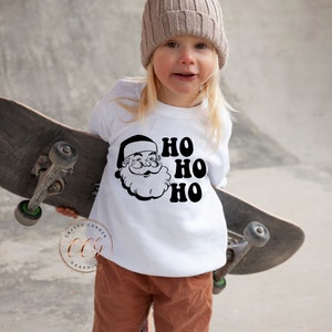 Peut inclure: Un t-shirt blanc &agrave; manches longues avec un motif noir et blanc de P&egrave;re No&euml;l et le texte "HO HO HO". Un enfant porte le t-shirt et tient une planche &agrave; roulettes.