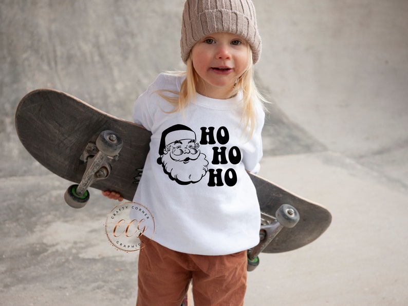 Peut inclure: Un t-shirt blanc &agrave; manches longues avec un motif noir et blanc de P&egrave;re No&euml;l et le texte "HO HO HO". Un enfant porte le t-shirt et tient une planche &agrave; roulettes.
