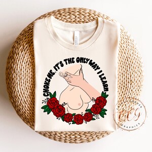 Puede incluir: Camiseta color crema con un dise&ntilde;o gr&aacute;fico. El dise&ntilde;o presenta una mano alrededor de un cuello, rodeada por una corona de rosas rojas y el texto "CHOKE ME. IT'S THE ONLY WAY I LEARN" en un c&iacute;rculo.