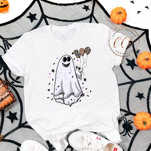 K&ouml;nnte beinhalten: Wei&szlig;es T-Shirt mit einem Cartoon-Geist, der Ballons h&auml;lt. Der Geist l&auml;chelt und hat einen schwarzen Umriss. Die Ballons sind orange, schwarz und lila. Um den Geist herum befinden sich schwarze Sterne und Konfetti.