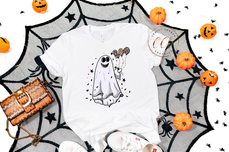 K&ouml;nnte beinhalten: Wei&szlig;es T-Shirt mit einem Cartoon-Geist, der Ballons h&auml;lt. Der Geist l&auml;chelt und hat einen schwarzen Umriss. Die Ballons sind orange, schwarz und lila. Um den Geist herum befinden sich schwarze Sterne und Konfetti.