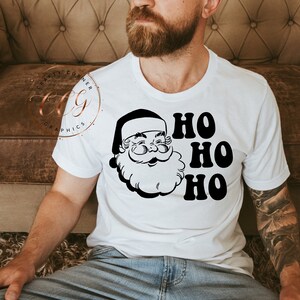 Peut inclure: T-shirt blanc avec une illustration en noir et blanc du visage du P&egrave;re No&euml;l et le texte "HO HO HO" en noir.