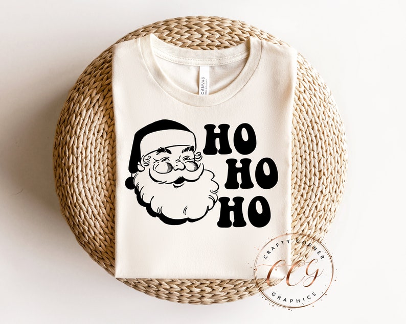 Peut inclure: Un t-shirt blanc avec une illustration en noir et blanc du P&egrave;re No&euml;l et le texte "Ho Ho Ho" dans une police r&eacute;tro. Le t-shirt a &eacute;galement un petit logo en bas &agrave; droite qui dit "Crafty Corner Graphics".