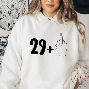 Puede incluir: Sudadera blanca con texto negro que dice "29+" y un contorno negro de un dedo medio.