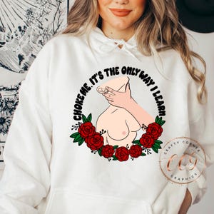 Puede incluir: Sudadera con capucha blanca con un dise&ntilde;o gr&aacute;fico. El dise&ntilde;o incluye una ilustraci&oacute;n estilizada de un torso con una mano alrededor del cuello, rodeado de rosas rojas y el texto "CHOKE ME, IT'S THE ONLY WAY I LEARN".